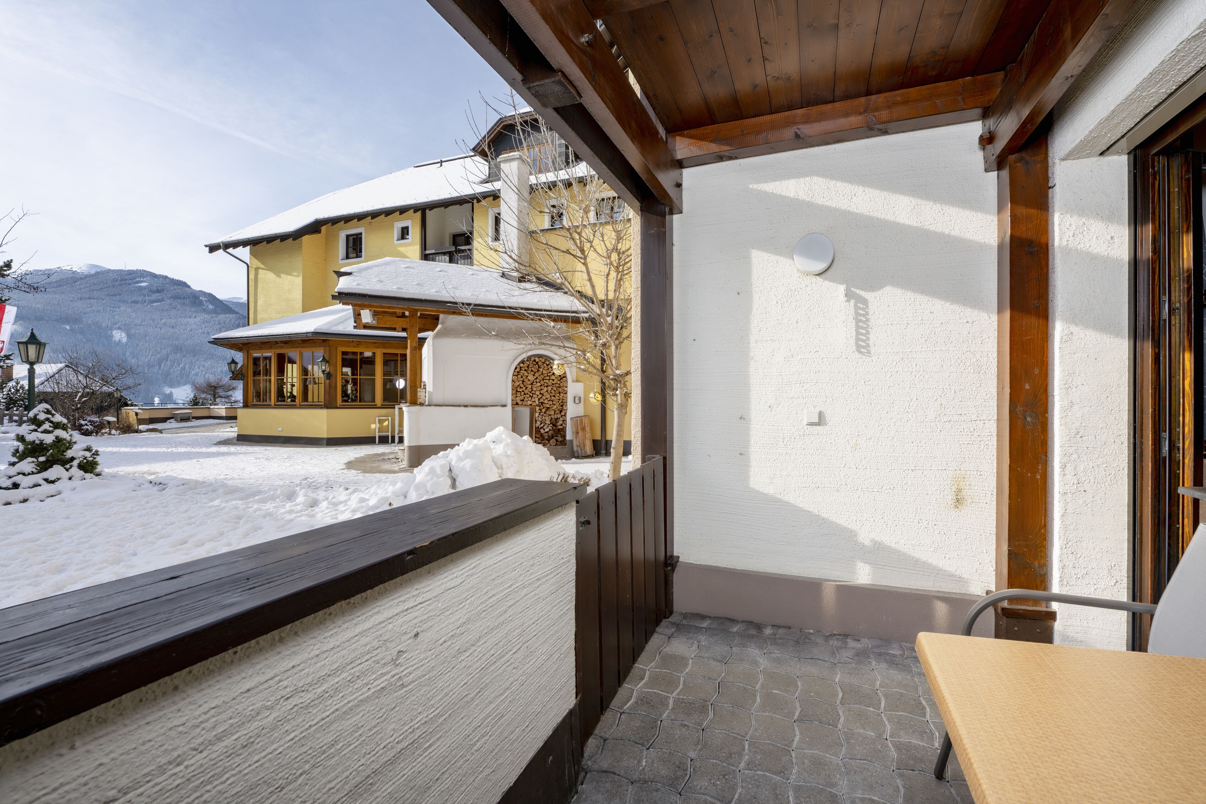A covered outdoor area with a wooden bench and a table, a snow-covered ground, and a building with a yellow facade and a white wall in the background.