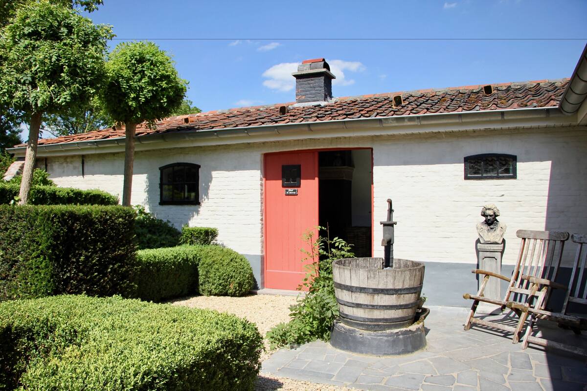 Un bâtiment blanc avec une porte rouge, un puits en pierre et une chaise en bois.