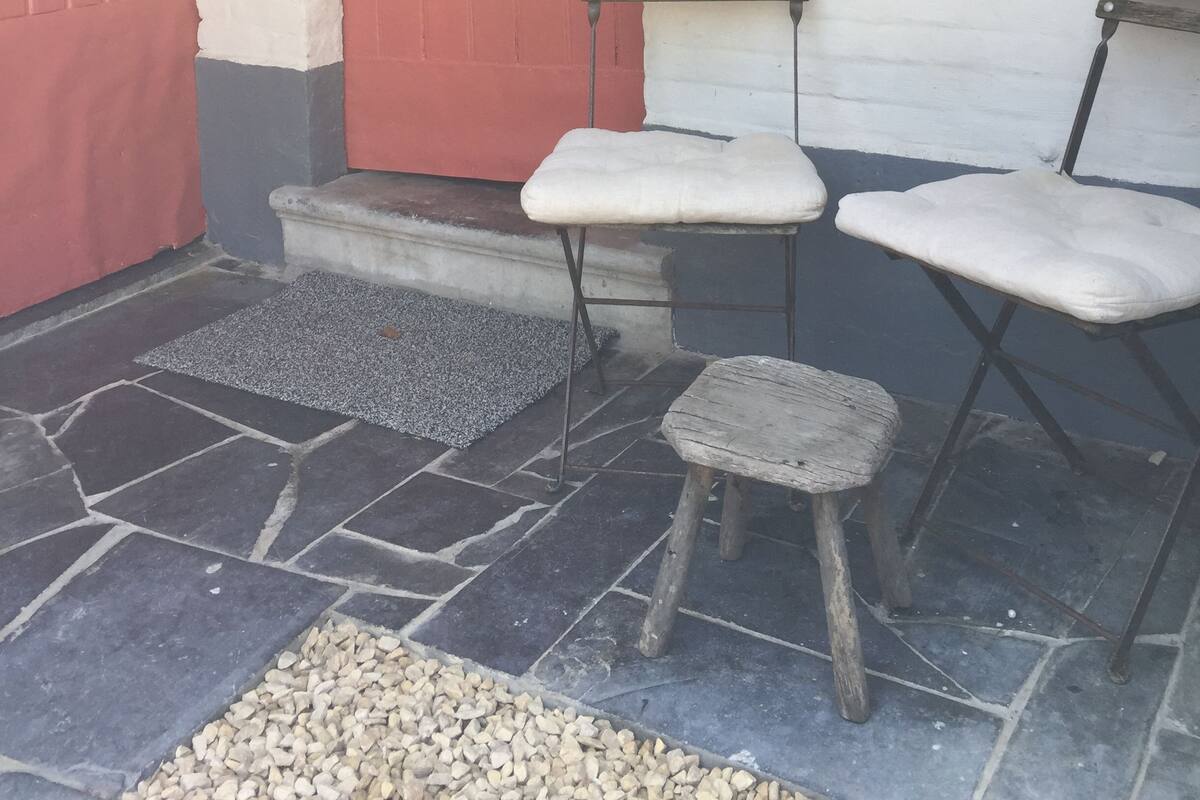 Un bâtiment avec une porte rouge et un mur blanc. On y voit deux chaises en bois avec des coussins, placées à l’extérieur.