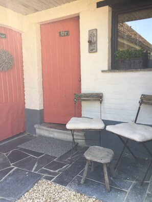 A building with a red door and a white wall. There are two wooden chairs with cushions outside. - B&B Het Stille Genoegen (Brakel)