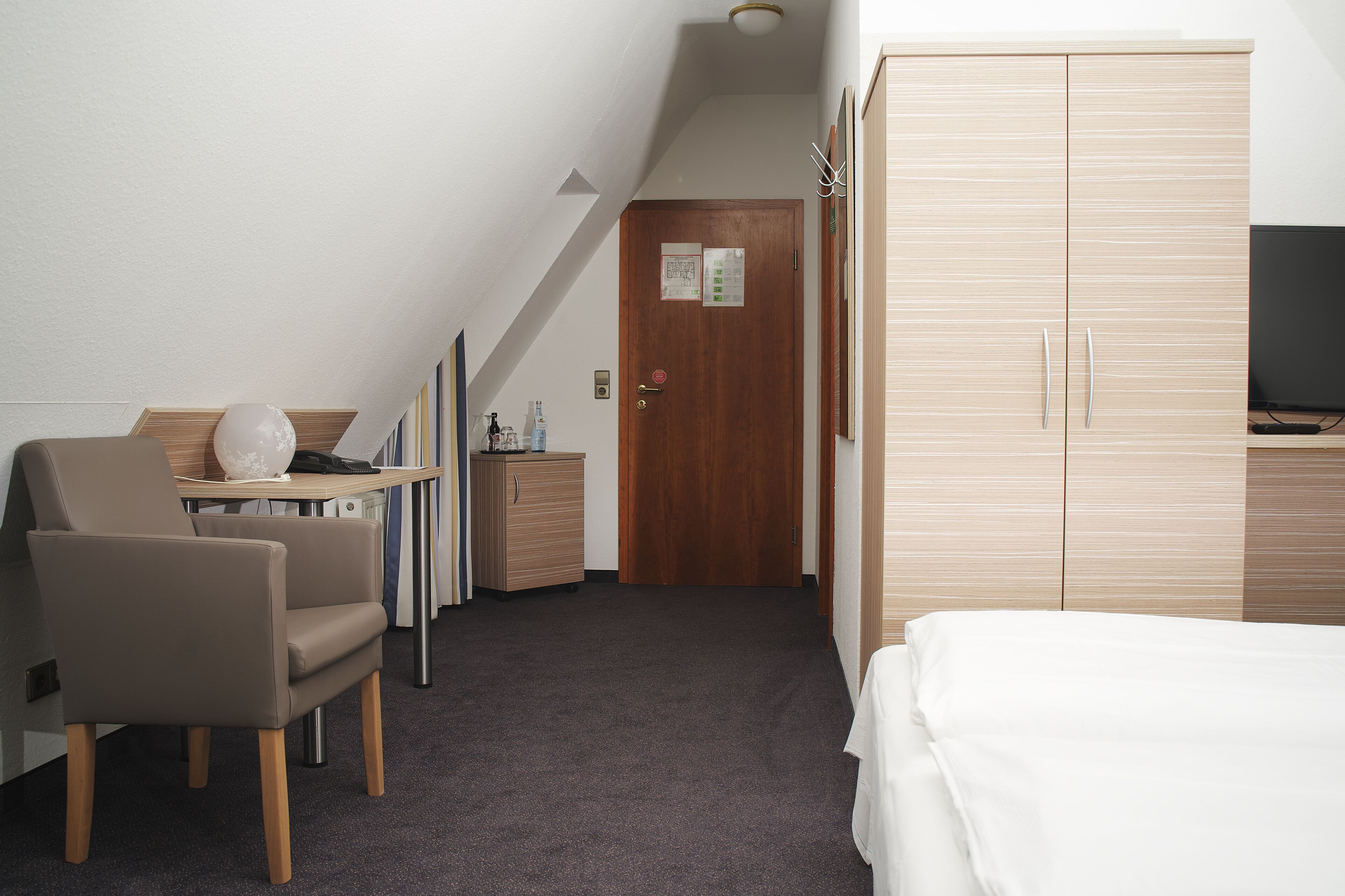 A hotel room with a bed, a desk with a chair, a wardrobe, and a television.