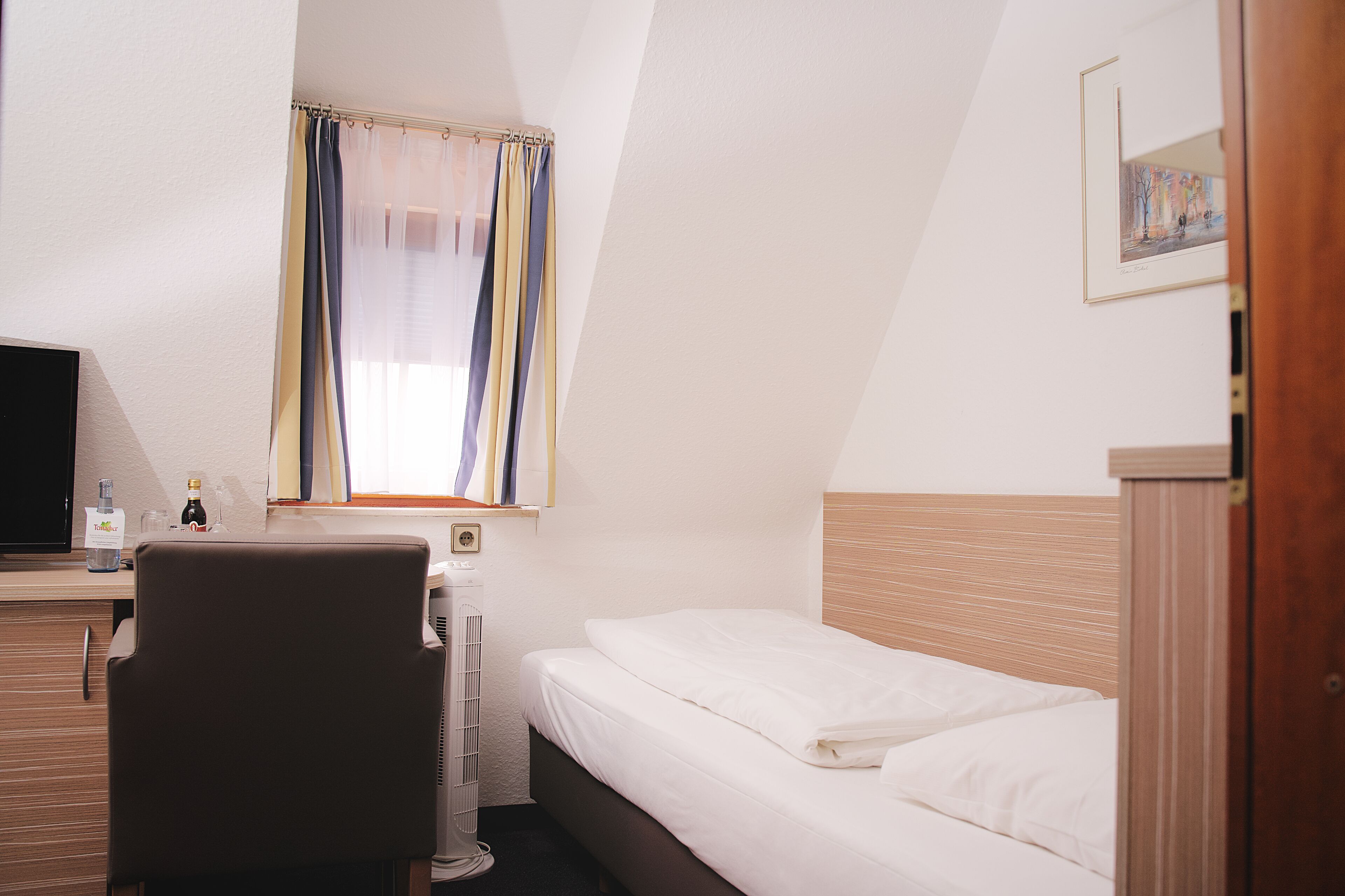 Une chambre d’hôtel avec un lit, un bureau avec un ordinateur, une chaise, une fenêtre avec des rideaux et un tableau encadré au mur.