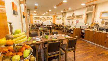 A restaurant with multiple dining tables and chairs, a buffet with fruits and pastries, and a kitchen area with shelves and appliances.