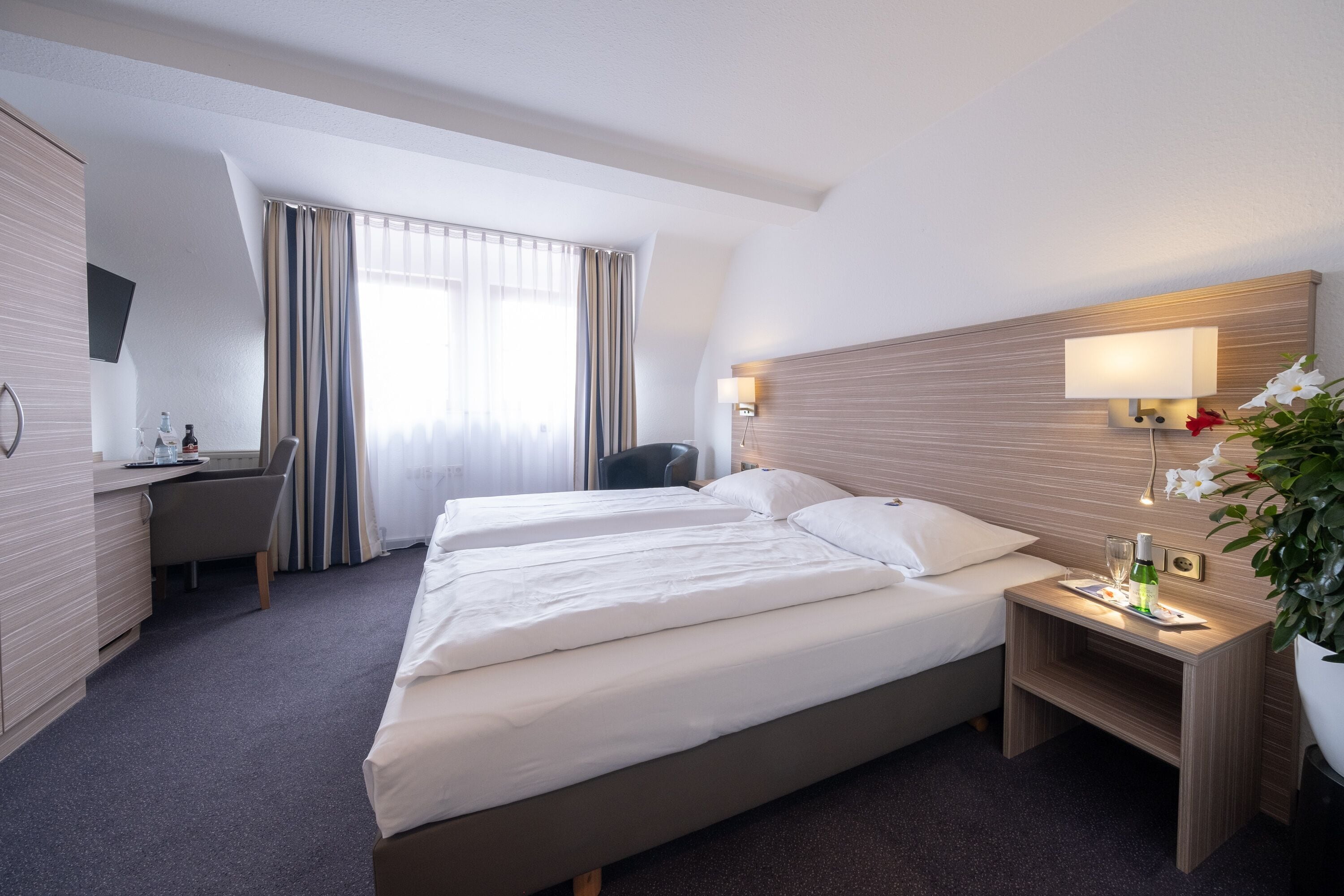 A hotel room with two beds, a desk, a chair, a bedside table with a lamp, and a large window with curtains.