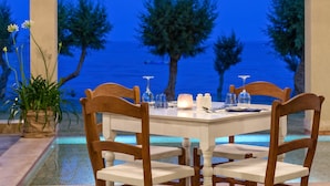 Un coin repas avec une table dressée pour quatre personnes, entourée de chaises en bois, donnant sur une piscine et offrant une vue sur la mer au crépuscule.