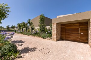 Uma entrada pavimentada conduz a uma garagem com design moderno, cercada por jardins bem cuidados e um céu azul límpido.