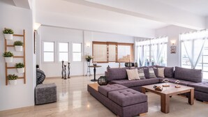 Una sala moderna con un sofá seccional, un reposapiés y una mesa de centro de madera. Hay plantas en macetas sobre una escalera de madera apoyada contra la pared, y una ventana grande con persianas.