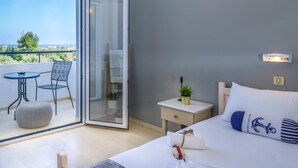 Habitación de hotel con una cama, una mesita de noche con lámpara, una silla, un otoman redondo y un balcón con mesa y sillas.
