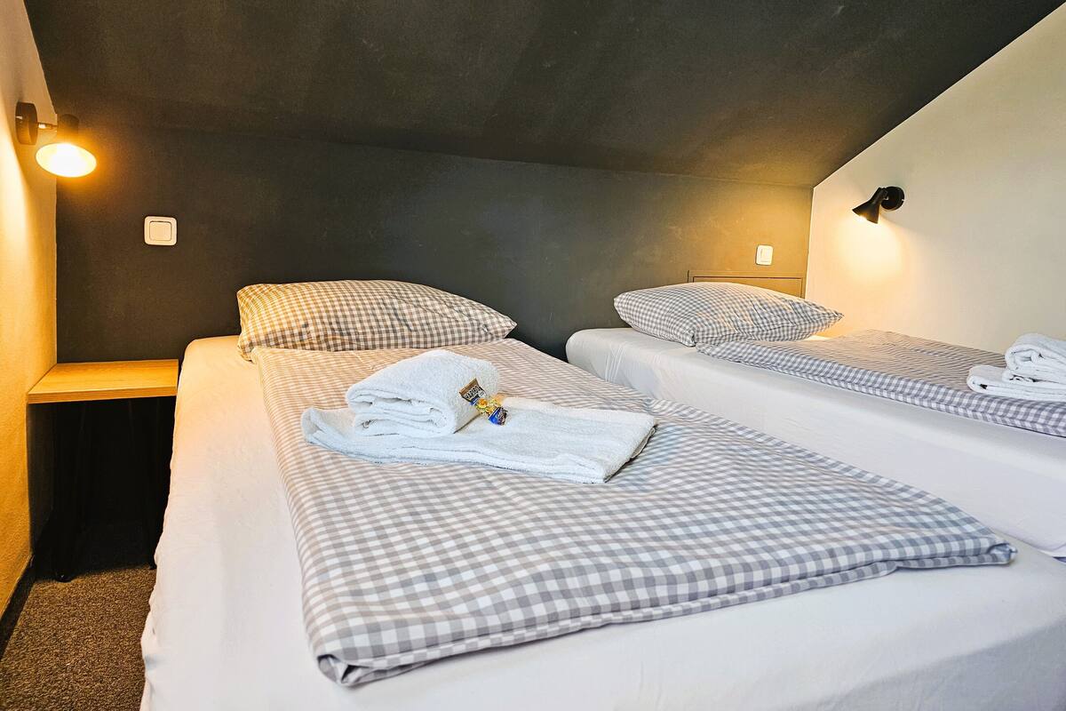 Een kamer met twee bedden, elk met geruit beddengoed en een opgevouwen handdoek op het bed. Aan weerszijden van de kamer zitten wandlampen.