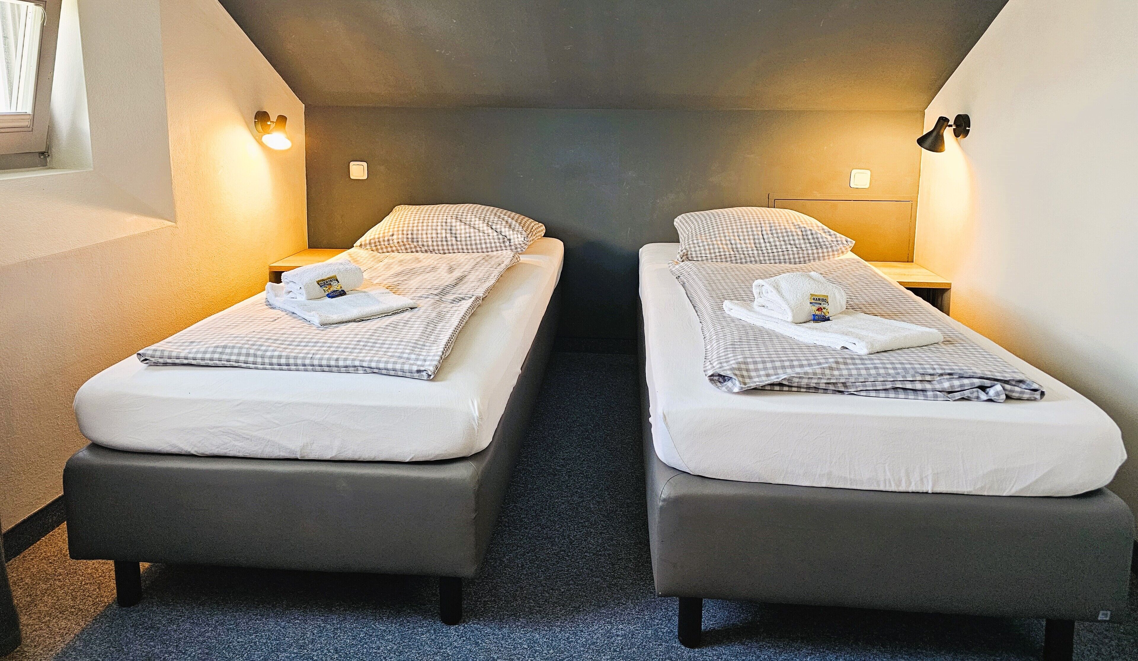 Due letti singoli con copriletto a quadretti, un comodino e una lampada fissata al muro.