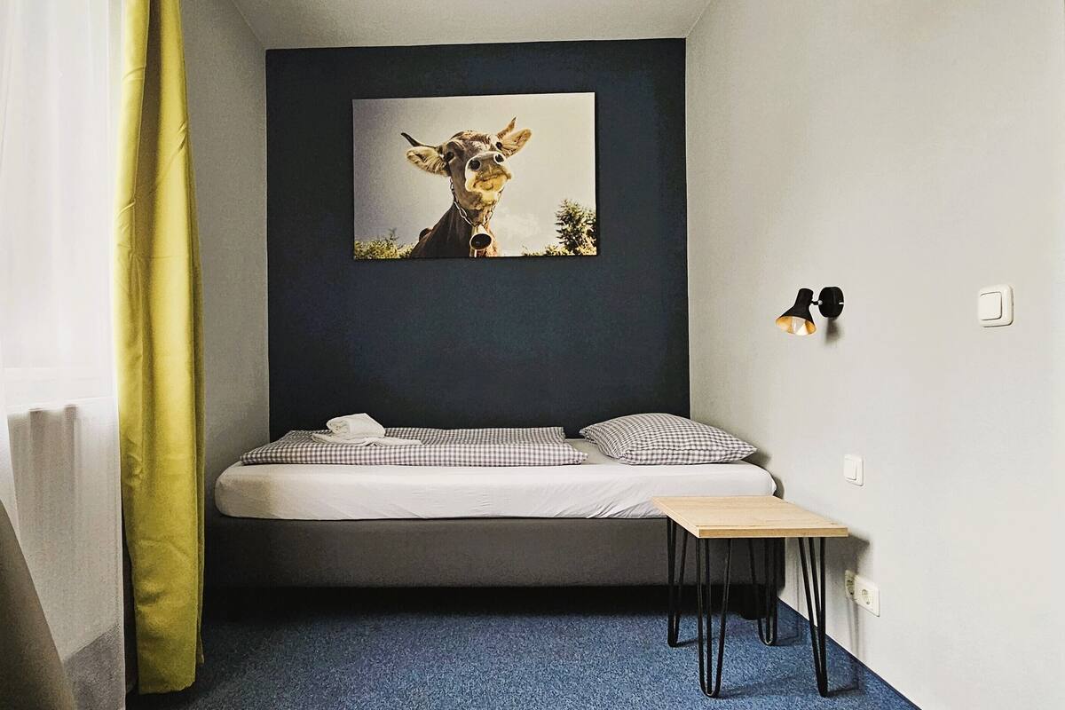 Een kleine kamer met een bed, een tafeltje, een aan de muur hangend beeld van een giraffe en een geel gordijn.