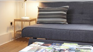 Een moderne slaapkamer met een grijze bank, een gestreept kussen, een nachtkastje met een lamp en een salontafel vol tijdschriften.