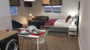 Een kleine hotelkamer met een bed, nachtkastje en een bescheiden kitchenette met een wasmachine.