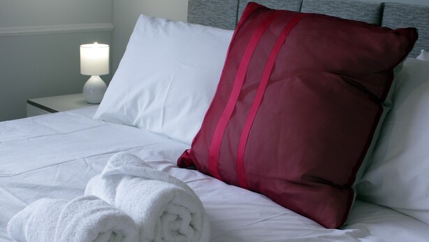 Een net opgemaakt bed met witte lakens, twee opgerolde witte handdoeken, een rood kussen en een wit kussen op een grijze hoofdbord.