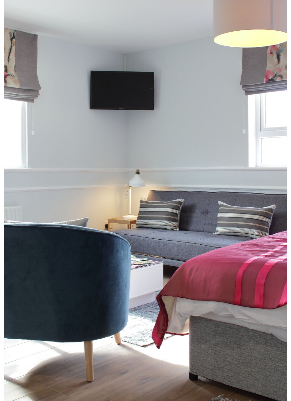 A modern bedroom with a grey sofa, a bed with a red blanket, a flat-screen TV mounted on the wall, and a lamp on a bedside table.