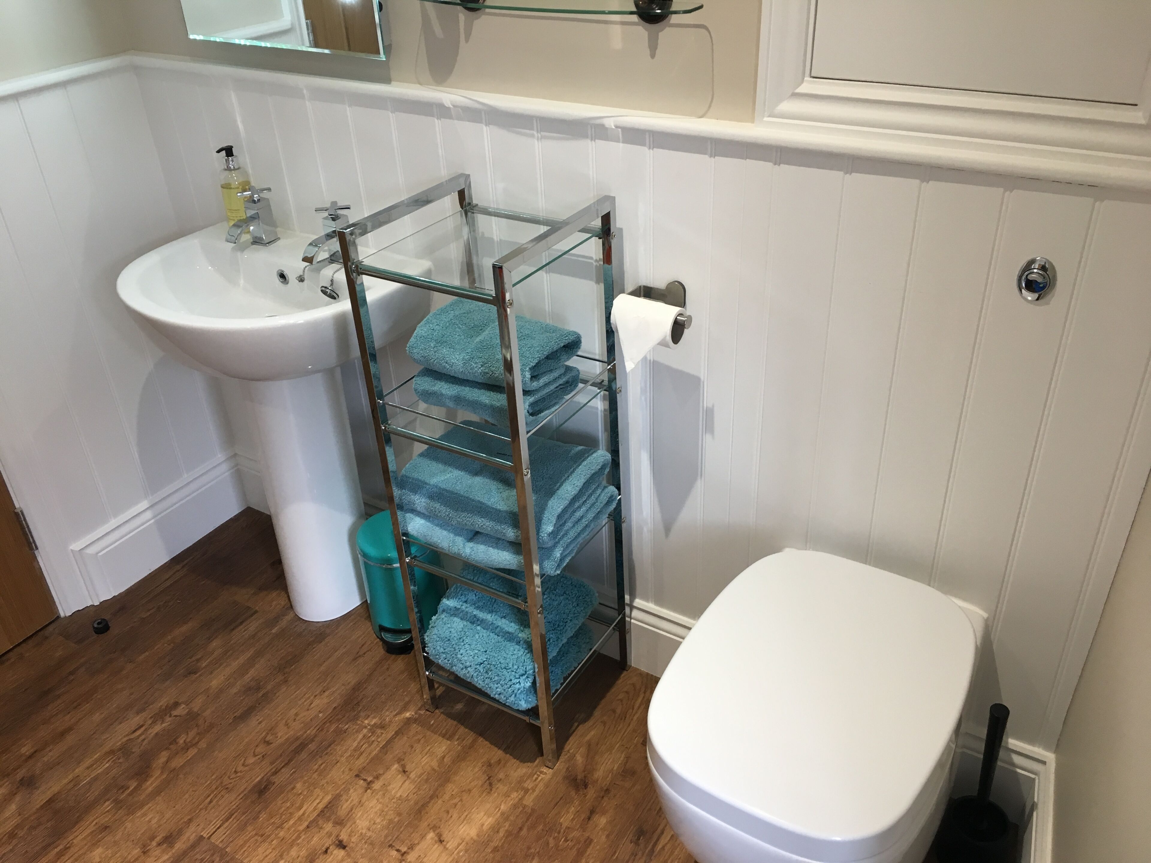 A bathroom with a white sink, a glass towel rack with folded towels, a toilet, and a trash bin.