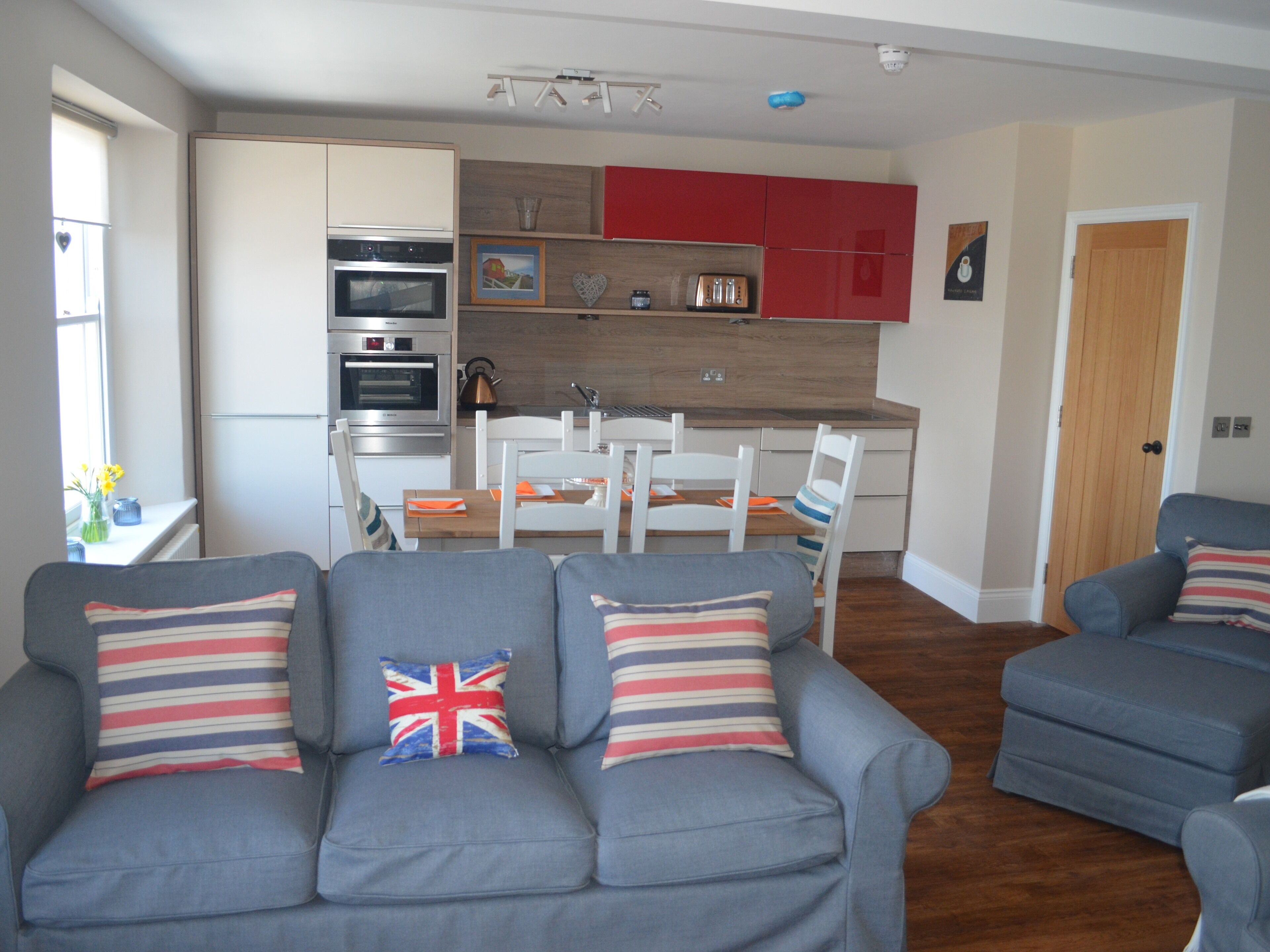A modern living room with grey sofas, a wooden dining table, and a kitchen with red cabinets and stainless steel appliances.