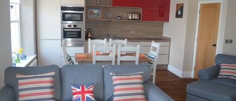 A modern living room with grey sofas, a wooden dining table, and a kitchen with red cabinets and stainless steel appliances.