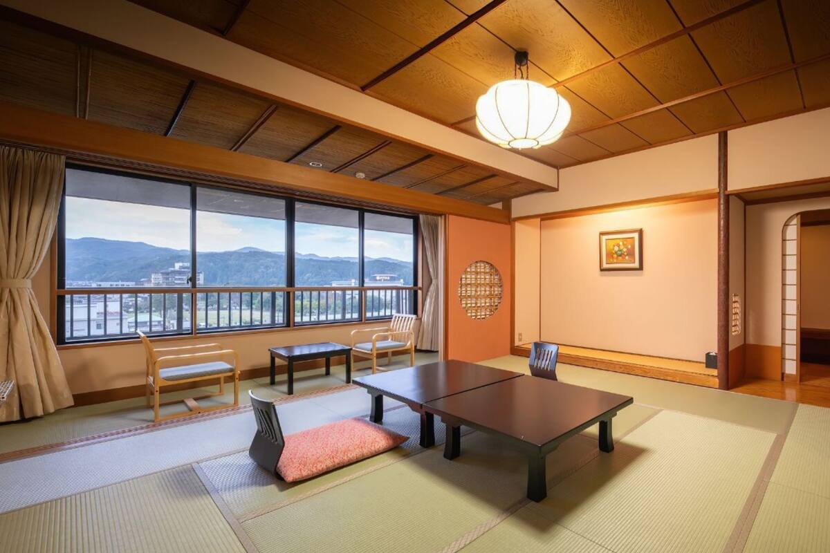 A traditional Japanese room with tatami flooring, a low table, a bench, and a view of mountains through large windows.