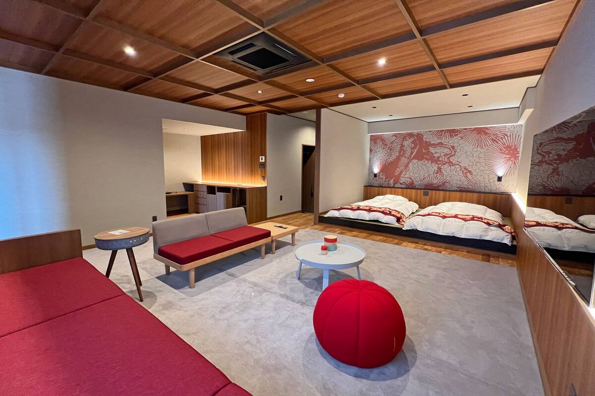 A modern hotel room with a wooden ceiling, a sofa, a bed with patterned bedding, a small round table, and a red pouf.