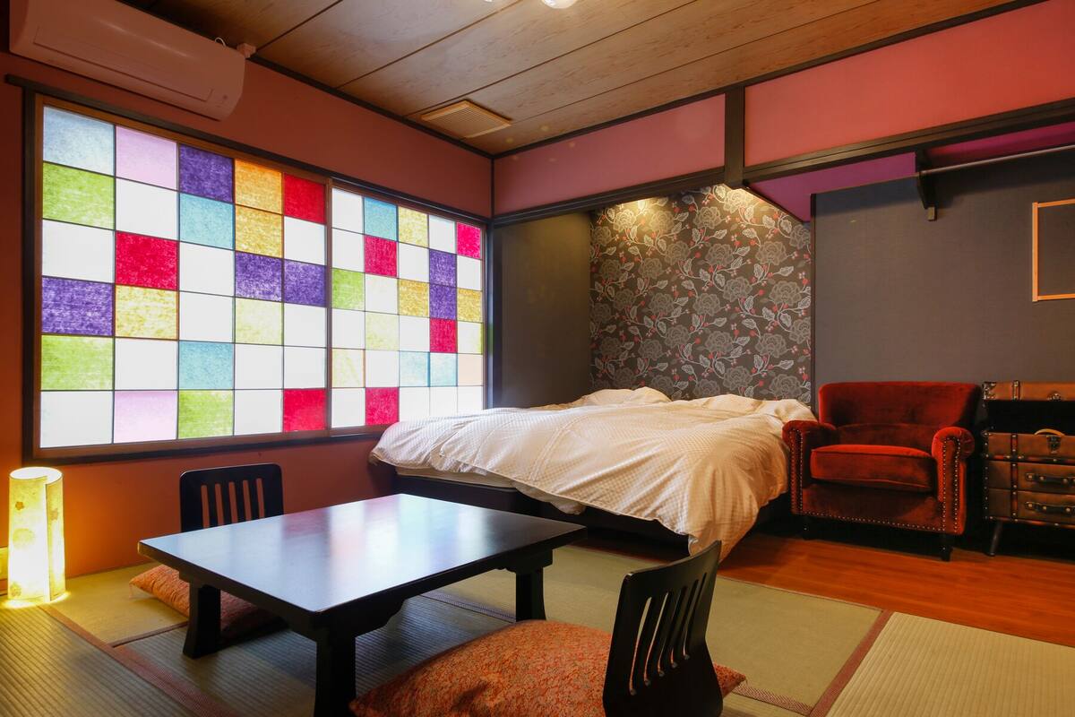 A room with a bed, a table, chairs, a red armchair, a suitcase, and a colorful stained glass window.