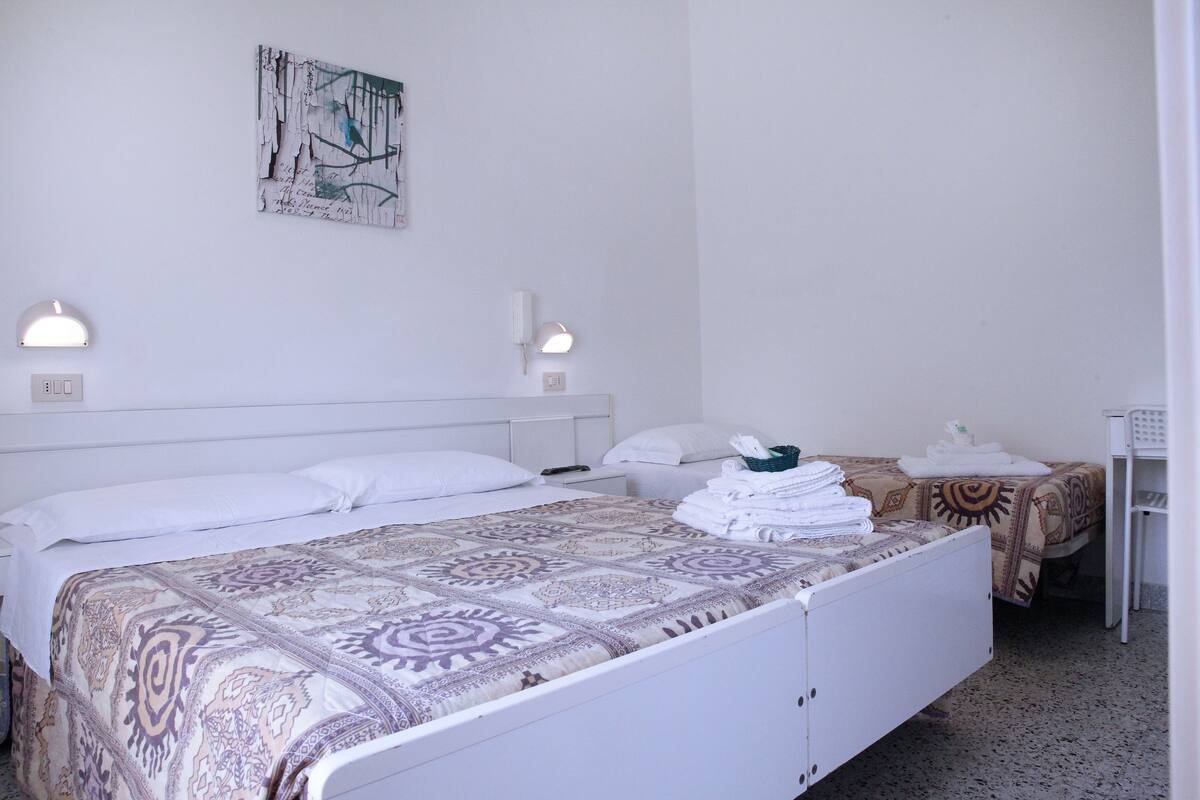 Une chambre d’hôtel propre et bien éclairée, avec un grand lit, deux tables de chevet, un petit bureau et une œuvre d’art accrochée au mur.