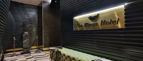 Un hall de hotel con un sofá tapizado de verde, una mesa de centro con estampado de mármol y un letrero que dice 'The Wings Hotel'.