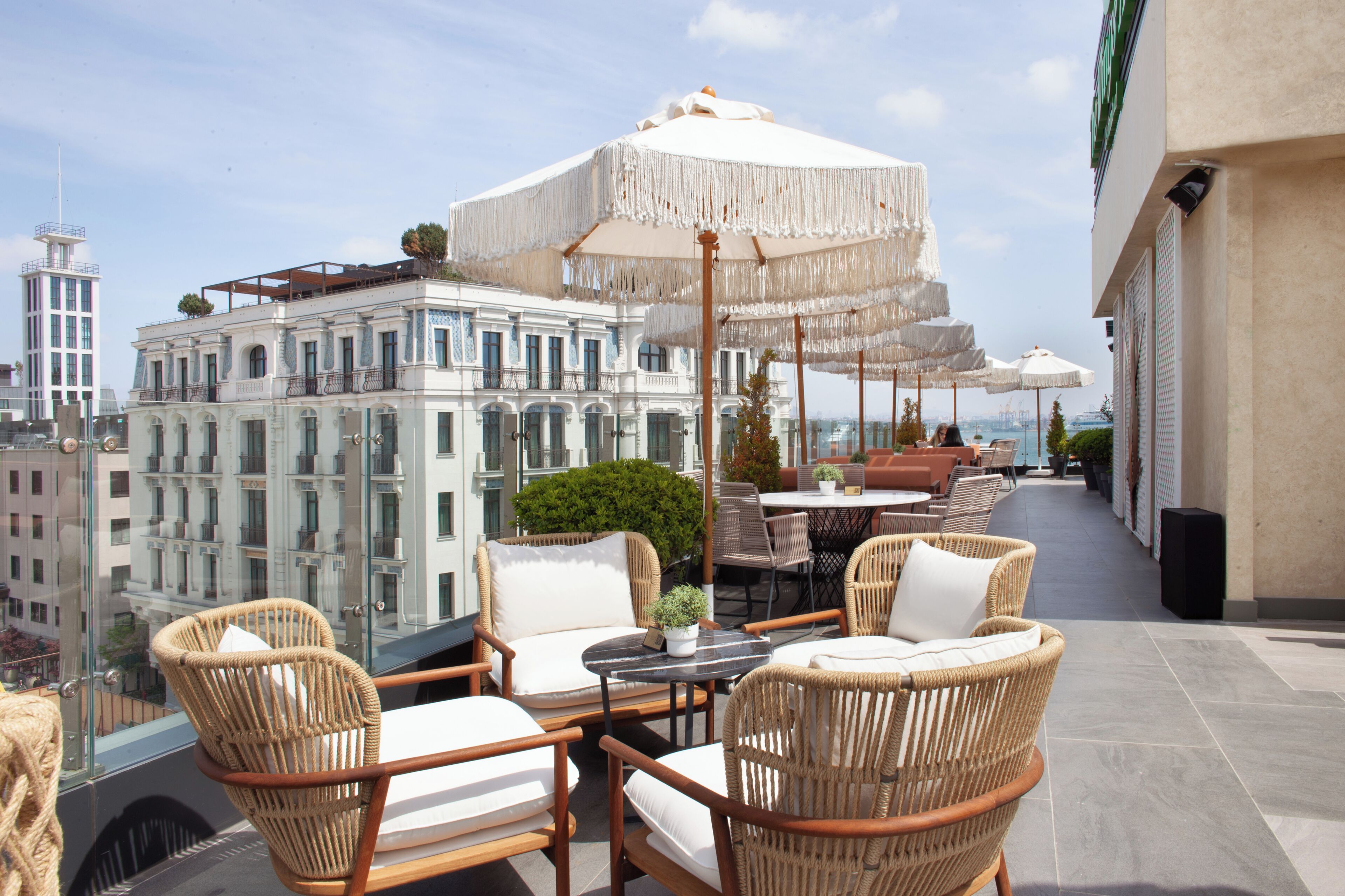 A rooftop terrace with wicker furniture, white umbrellas, and a view of the cityscape.