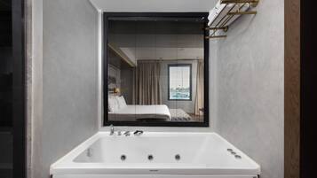 A modern bathroom with a large bathtub, a mirror, and a window with blinds.