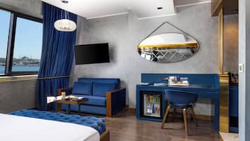 A modern hotel room with a bed, a sofa, a desk, and a chair. There is a TV mounted on the wall, a window with curtains, and a view of a cityscape.