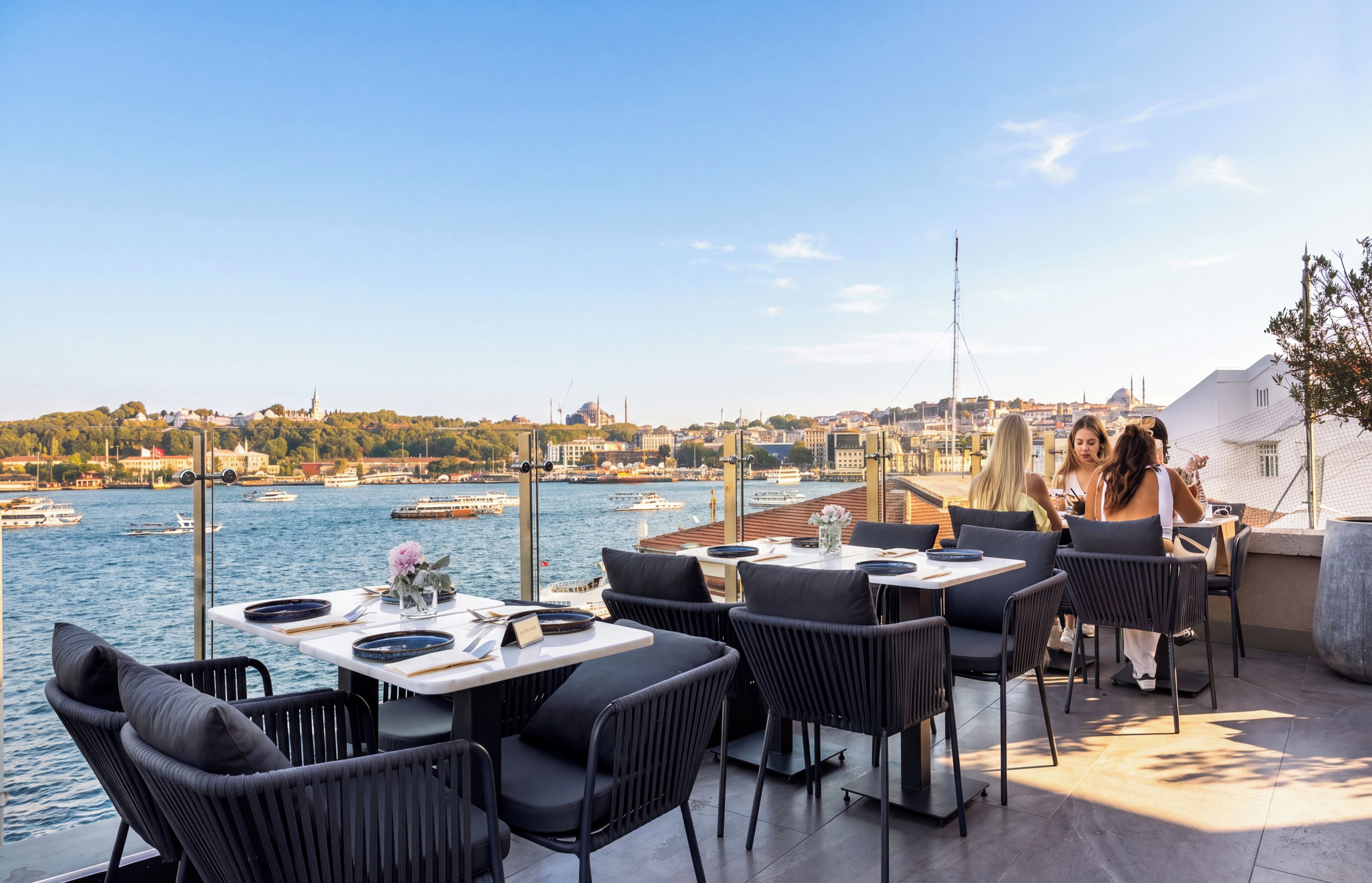 An outdoor dining area with tables and chairs overlooking a body of water and a cityscape.
