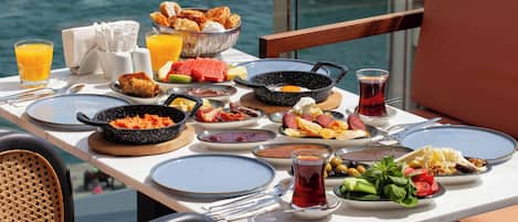 Mesa lista para el desayuno, con huevos, bollería y fruta, con vista a un cuerpo de agua.
