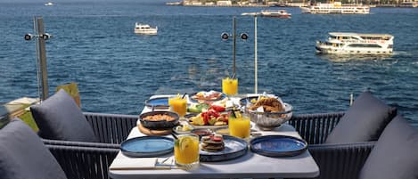 Mesa dispuesta para el desayuno con vista a un cuerpo de agua, barcos y un paisaje urbano distante.