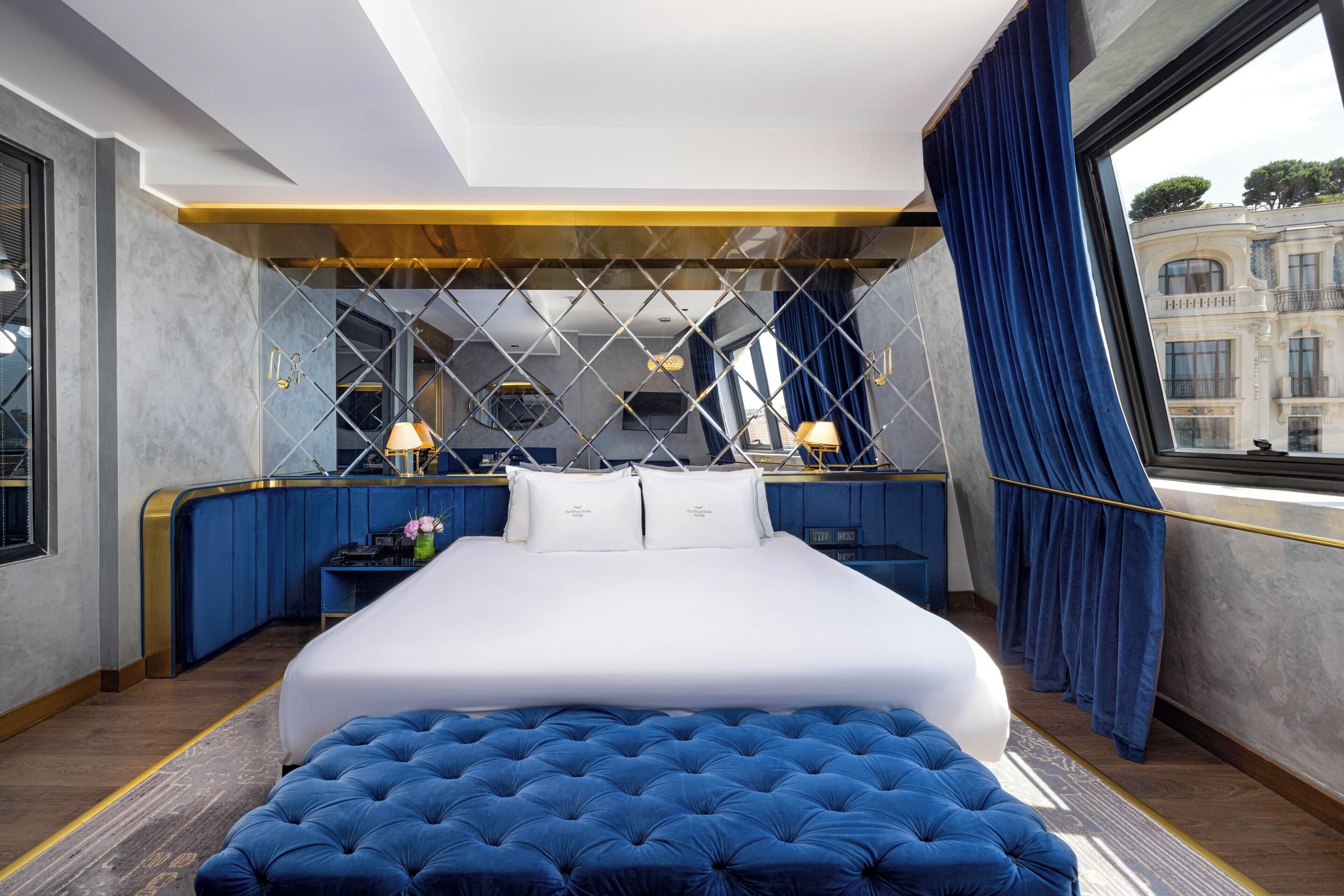 Habitación de hotel moderna con una cama grande, un banco tapizado en azul, un escritorio con lámpara y vistas a un edificio a través de una ventana.