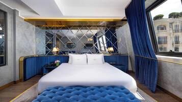 Habitación de hotel moderna con una cama grande, un banco tapizado en azul, un escritorio con lámpara y vistas a un edificio a través de una ventana.
