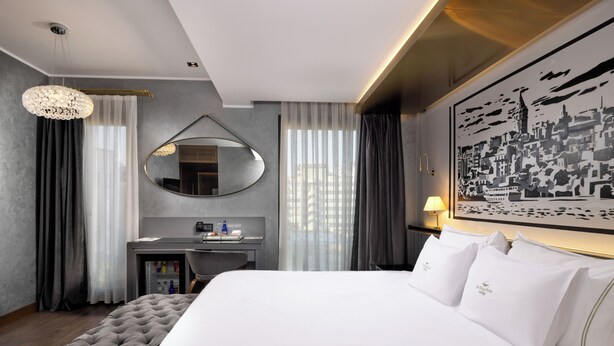 Habitación de hotel con una cama grande, un escritorio con espejo, un refrigerador pequeño y un cuadro de un paisaje urbano en la pared.