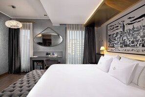 A hotel room with a large bed, a desk with a mirror, a small refrigerator, and a cityscape painting on the wall. - The Wings Hotels Karakoy (Istanbul)