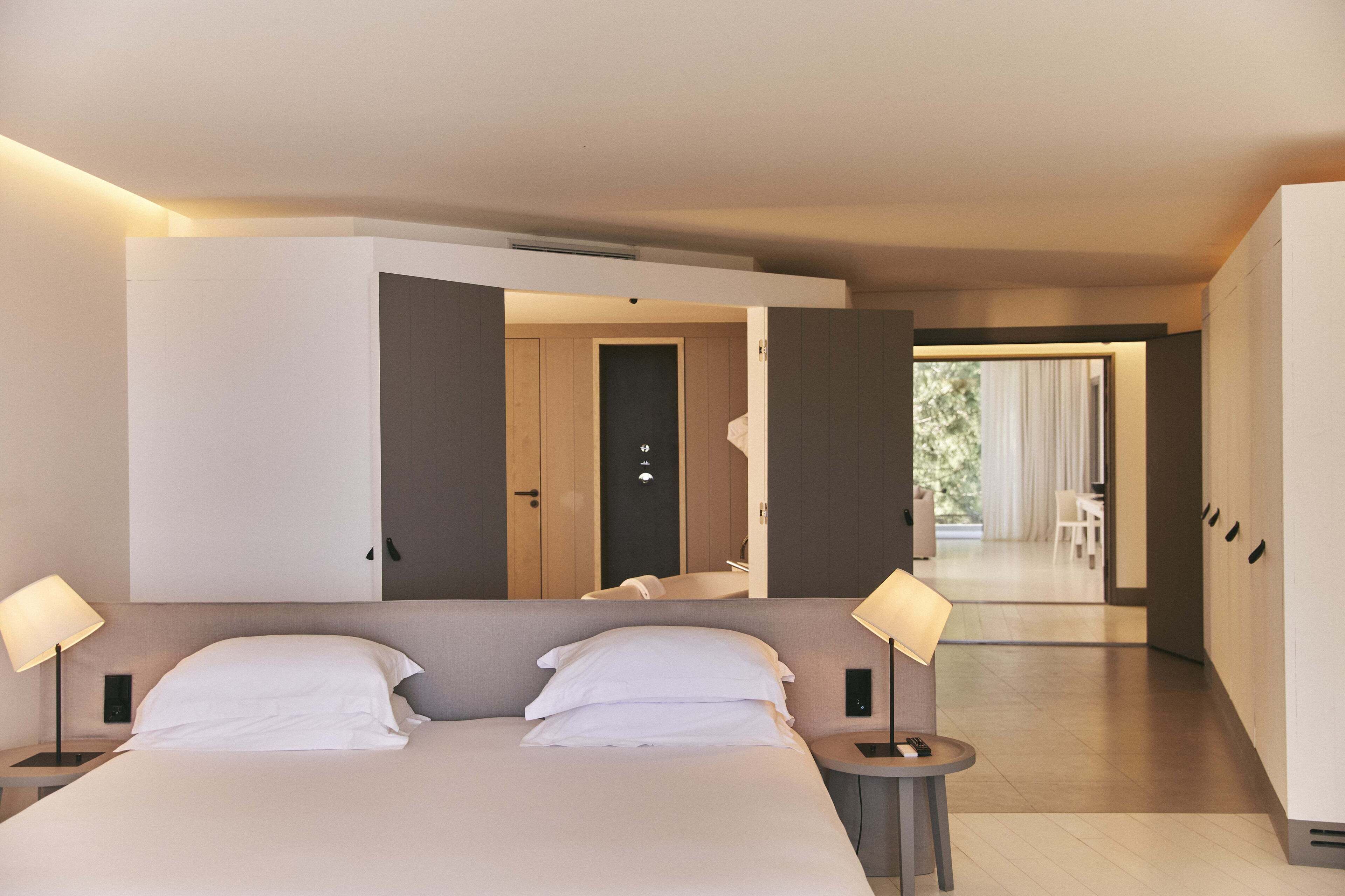 Une chambre d’hôtel moderne avec un grand lit, deux lampes de chevet, une petite table et une vue sur la salle à manger par une porte ouverte.