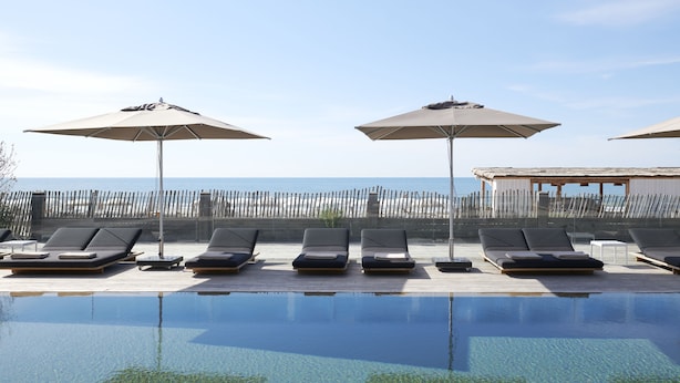 A poolside area with lounge chairs and umbrellas, a wooden deck, and a view of the sea. - Hôtel Plage Palace & Spa (Palavas-les-Flots)