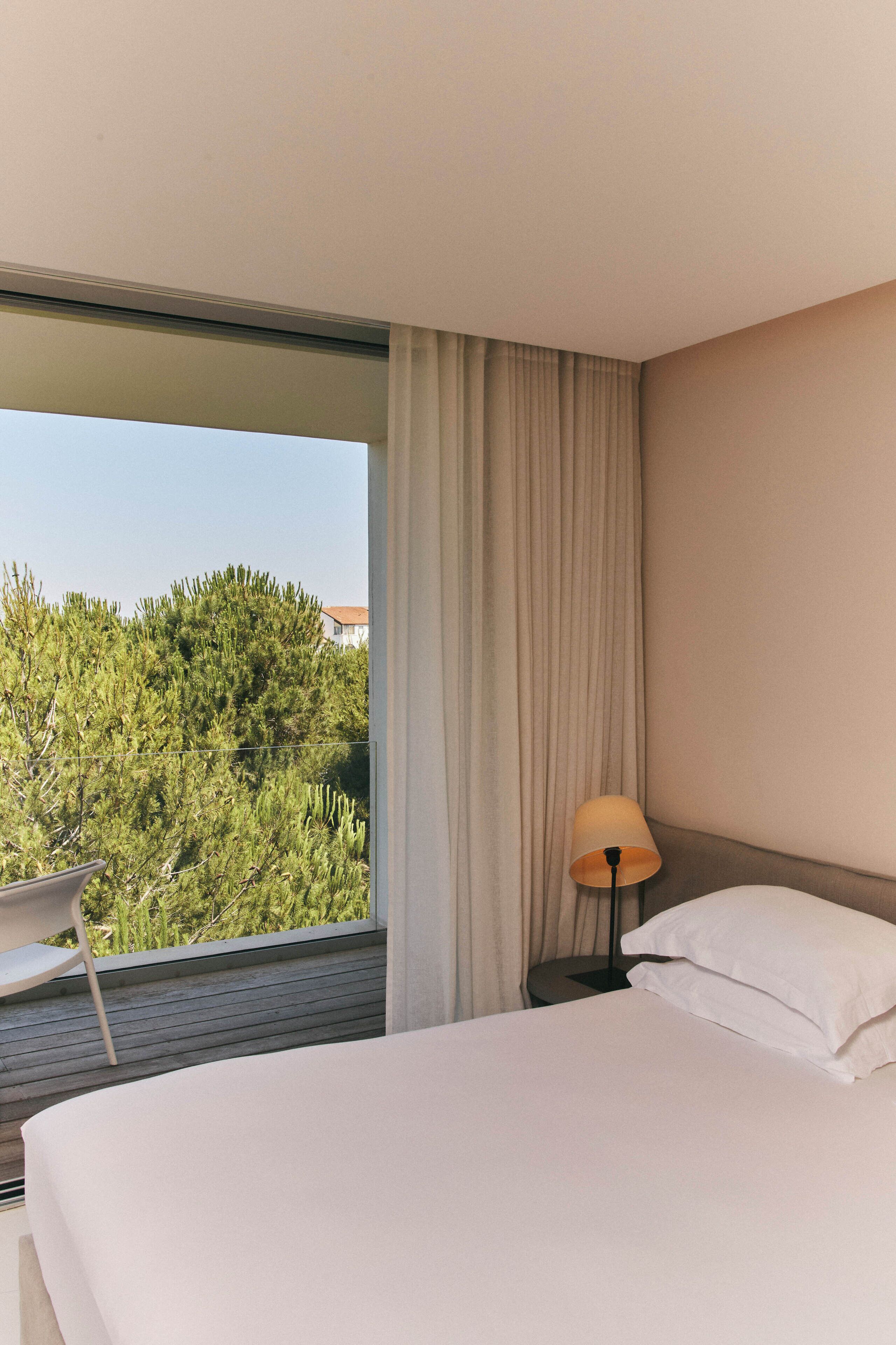 A hotel room with a large bed, a nightstand with a lamp, a chair, and a view of greenery through a window.
