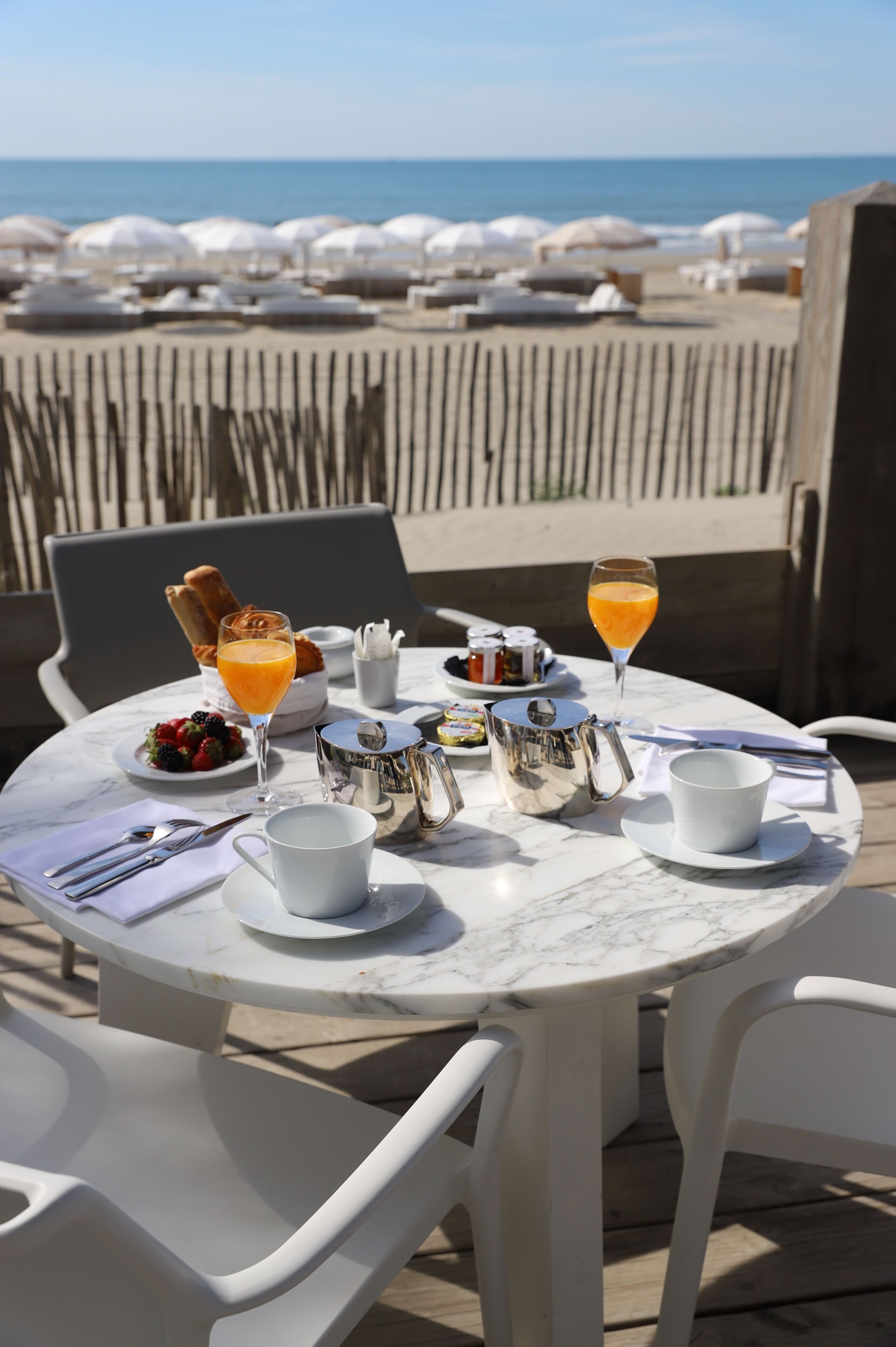 Une table ronde en marbre, dressée pour le petit-déjeuner, avec vue sur la plage et des parasols en arrière-plan.