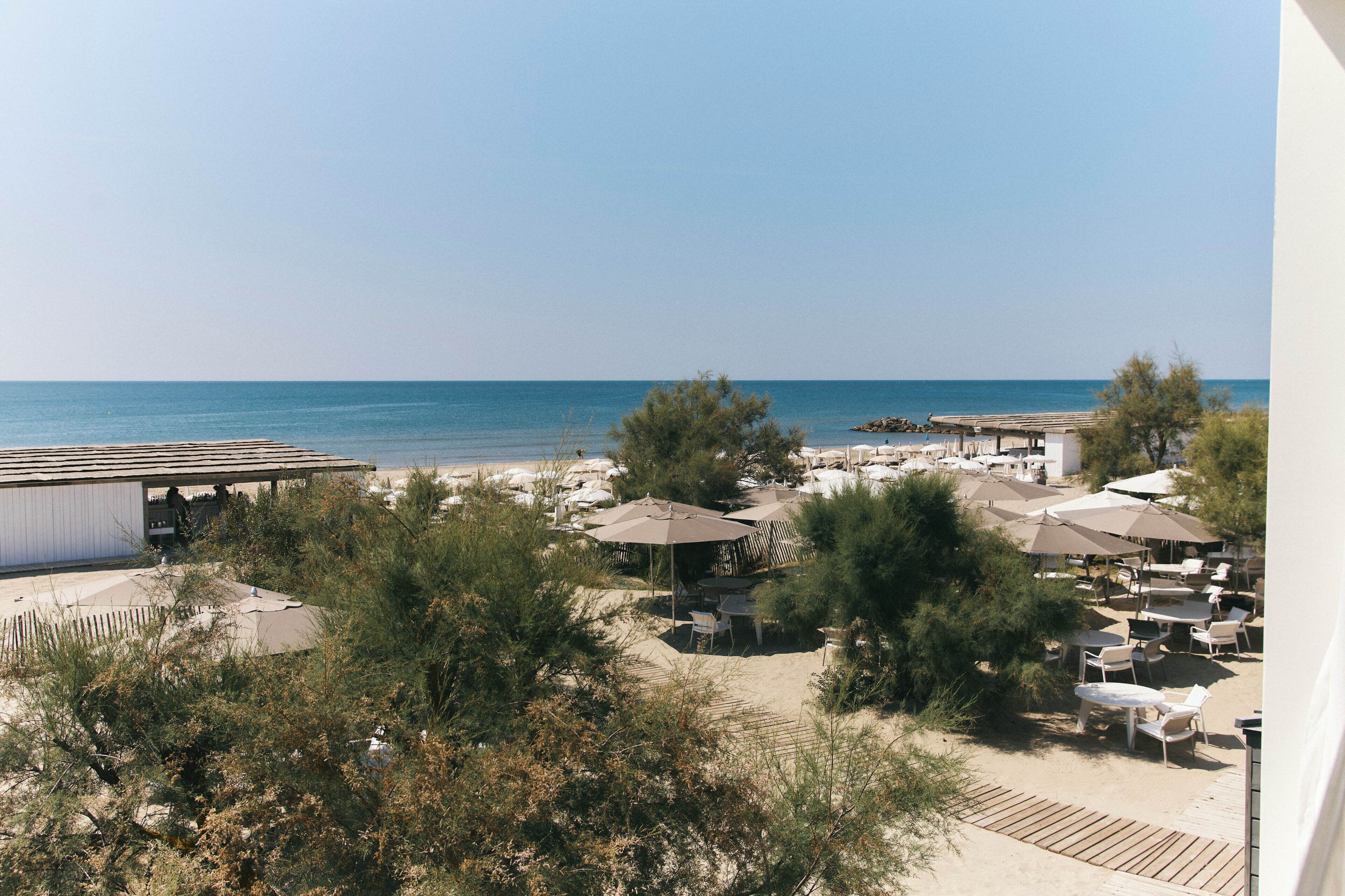 A beachside view with outdoor seating and umbrellas, a clear sky, and a calm sea.
