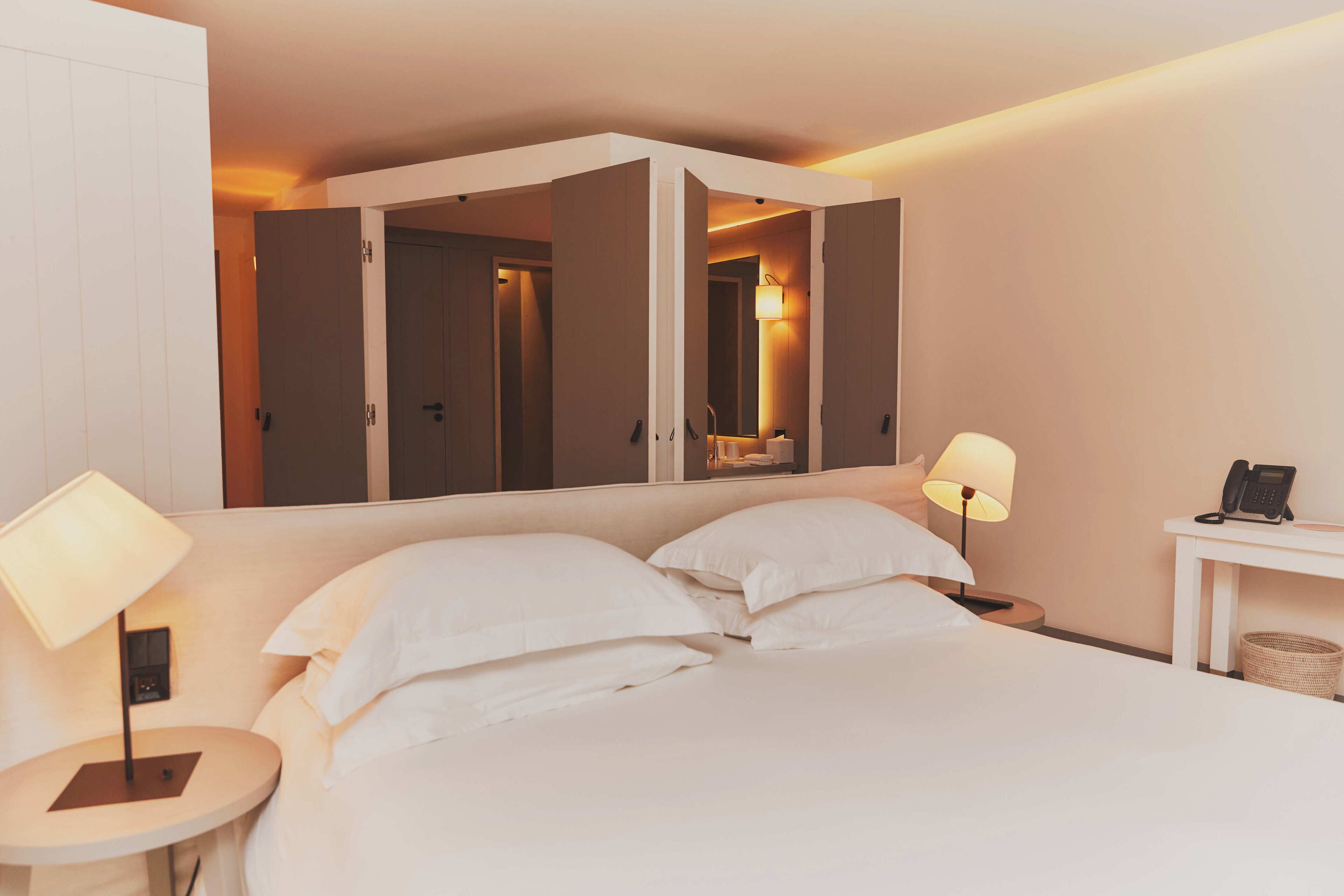 A hotel room with a large bed, two bedside tables with lamps, a small table with a phone, and a wardrobe with a mirror.