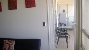 Ein Wohnzimmer mit einem dunkelgrauen Sofa und einem gemusterten Zierkissen, eine Glastür, die in einen Essbereich mit Tisch und Stühlen führt, und ein Kühlschrank im Hintergrund.