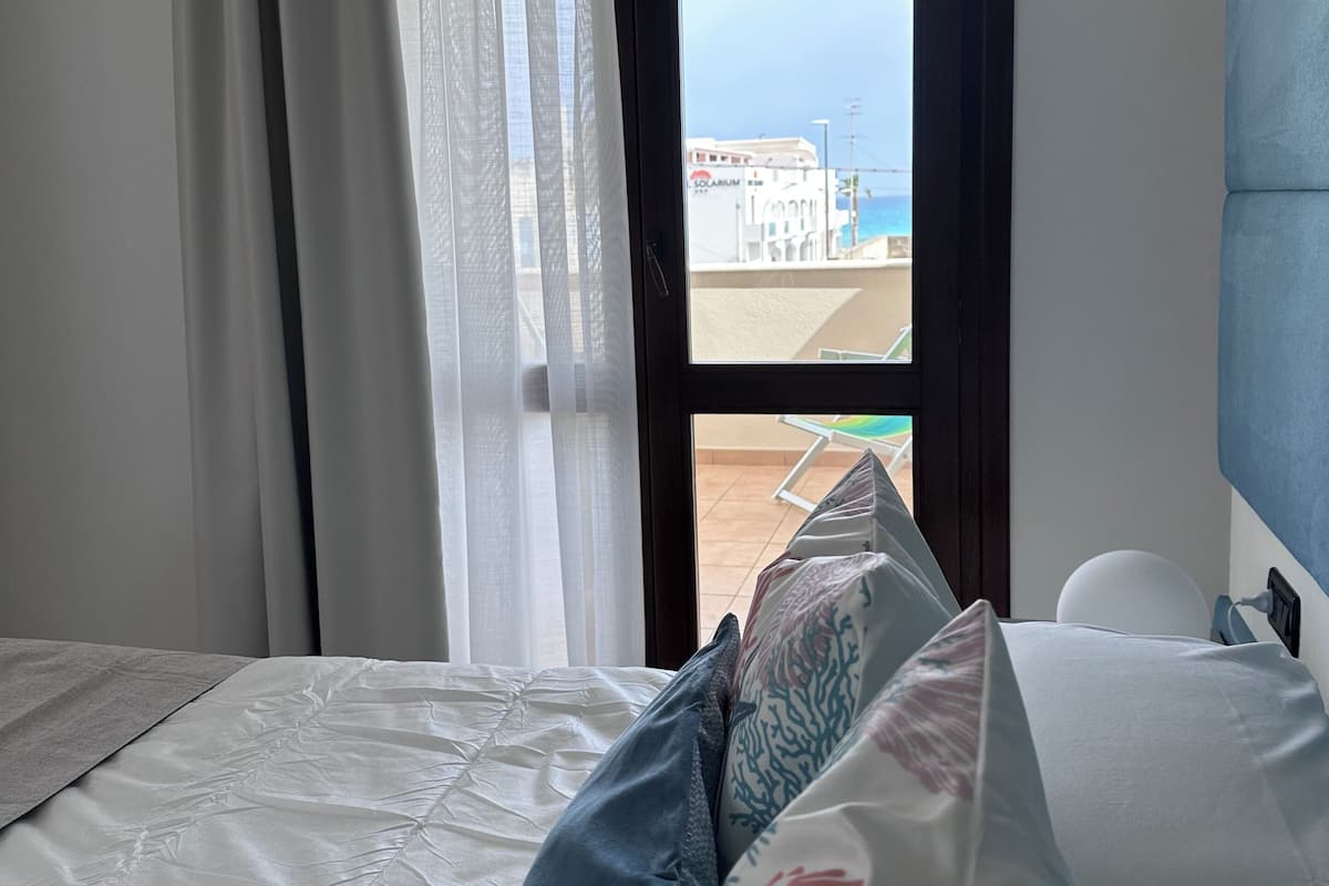 Une chambre avec un lit, une fenêtre avec des rideaux et une vue sur la mer et les bâtiments.