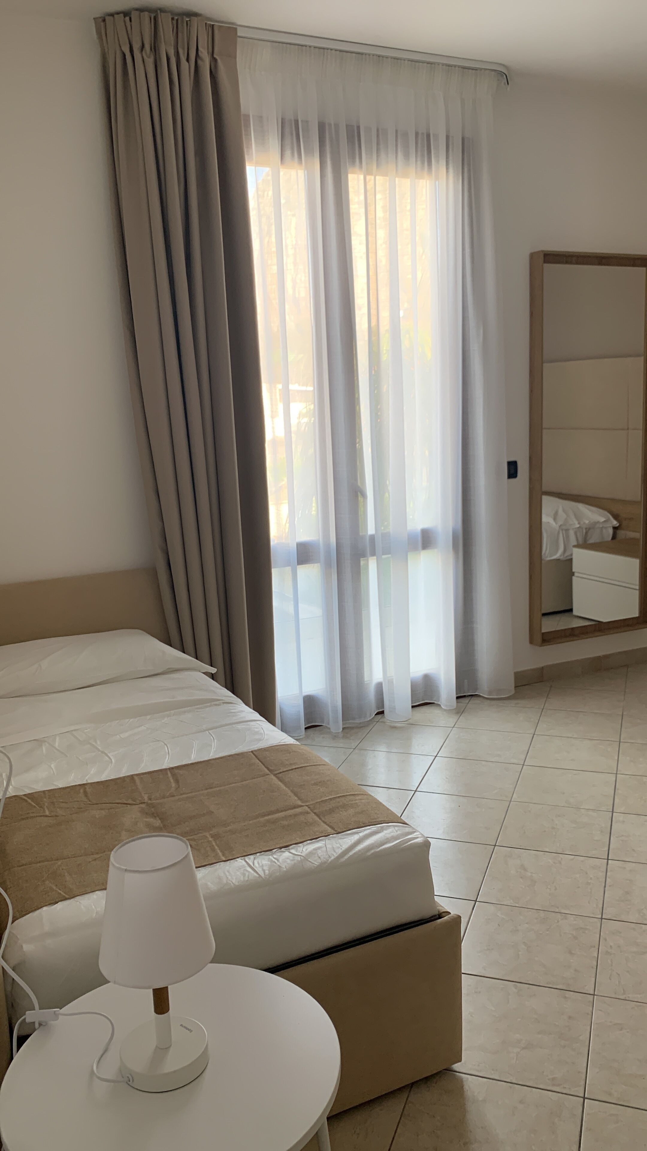 Ein Hotelzimmer mit Bett, Nachttisch mit Lampe, großem Fenster mit durchsichtigen Vorhängen und Kleiderschrank.