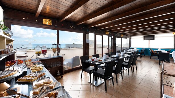 Un restaurant situé en bord de mer, avec des poutres en bois au plafond, des tables en plein air et un buffet proposant des pâtisseries et du pain.