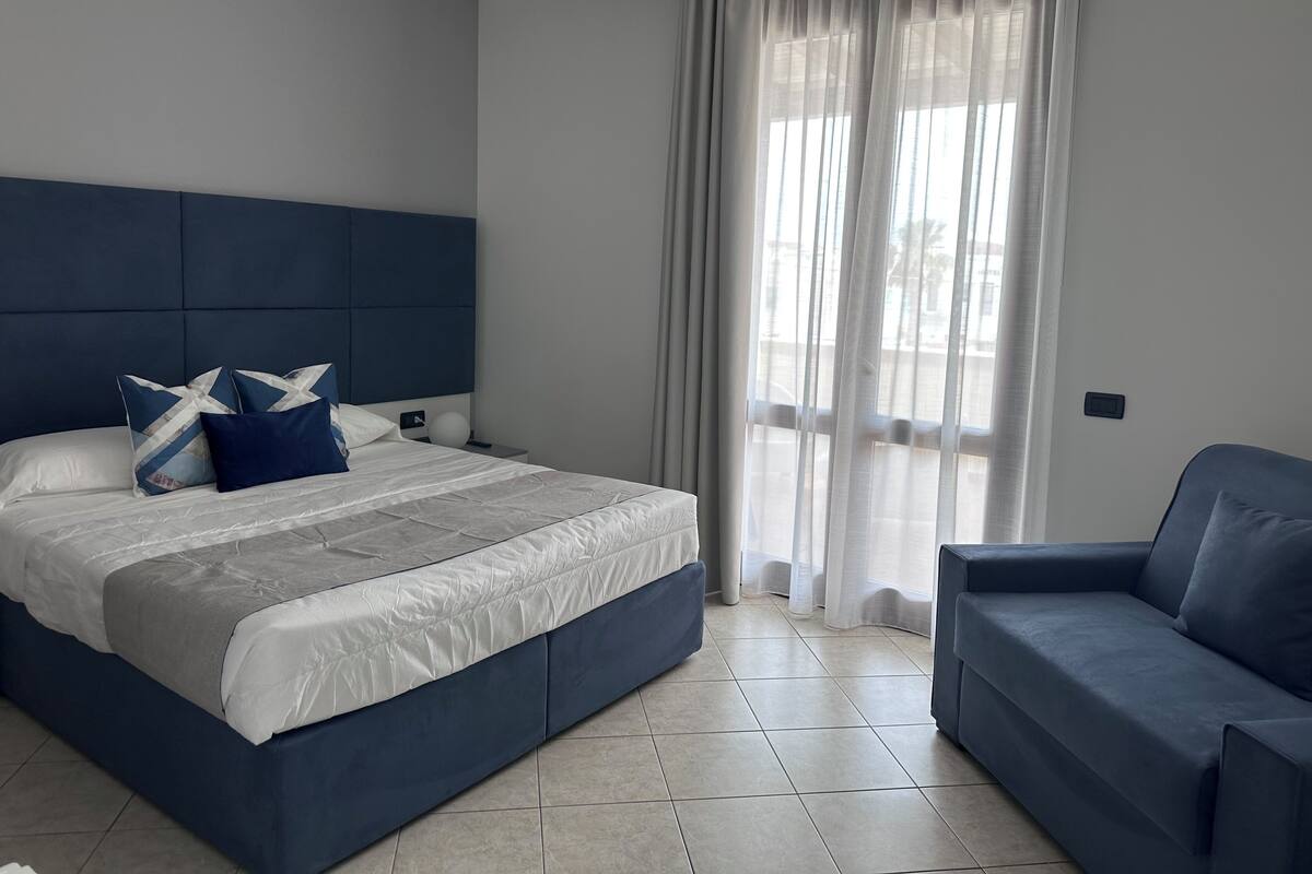 Une chambre à coucher moderne avec un grand lit, un canapé bleu et une vue sur l’extérieur à travers des rideaux transparents.