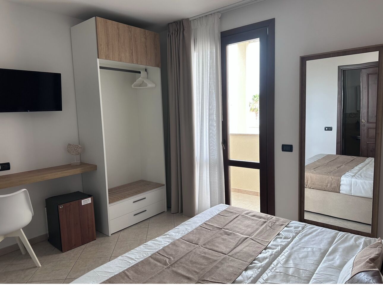 Una habitación de hotel con cama, escritorio, armario, televisor y balcón.