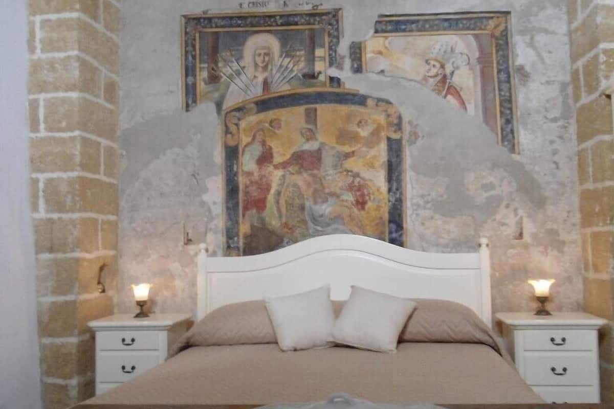 Un dormitorio con una cama, dos mesitas de noche, un muro decorativo con frescos, y una bandeja de desayuno con pasteles y tazas.